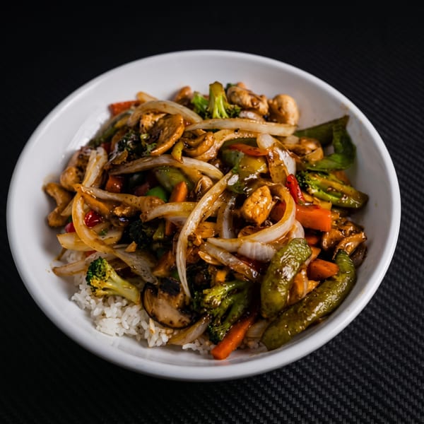 A white bowl filled with chicken stir-fry, rice, broccoli, onions, carrots, and mushrooms.