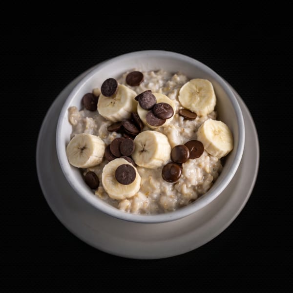 A white bowl of oatmeal topped with sliced bananas and chocolate chips on a dark background.