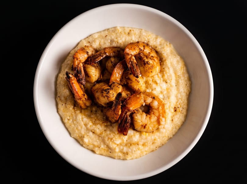 A white bowl filled with creamy grits topped with several seasoned, grilled shrimp.