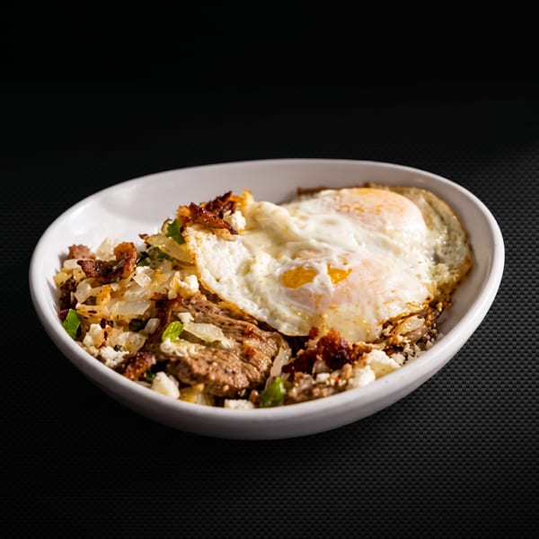 A white bowl filled with steak, onions, feta cheese, and two fried eggs on a dark background.